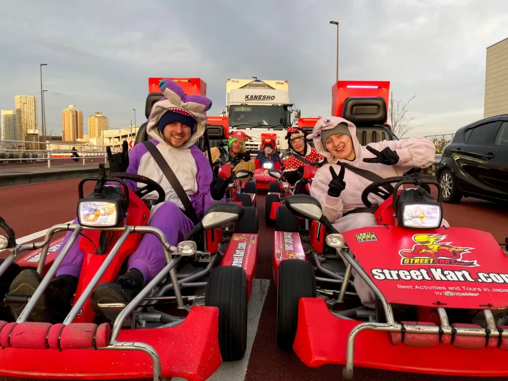 Group of people in colorful mascot costumes riding red go-karts on a city street, smiling and flashing peace signs at the camera.