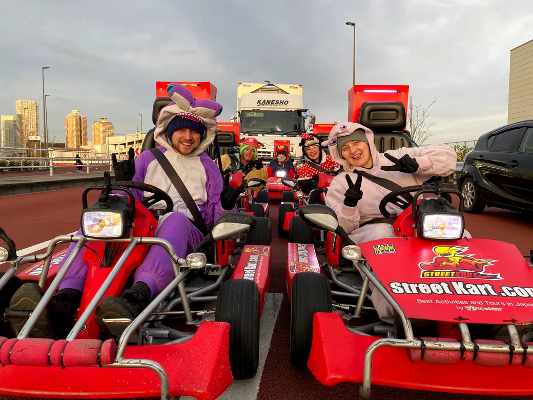Group of people in colorful animal onesies driving red go-karts on a track, waving at the camera.