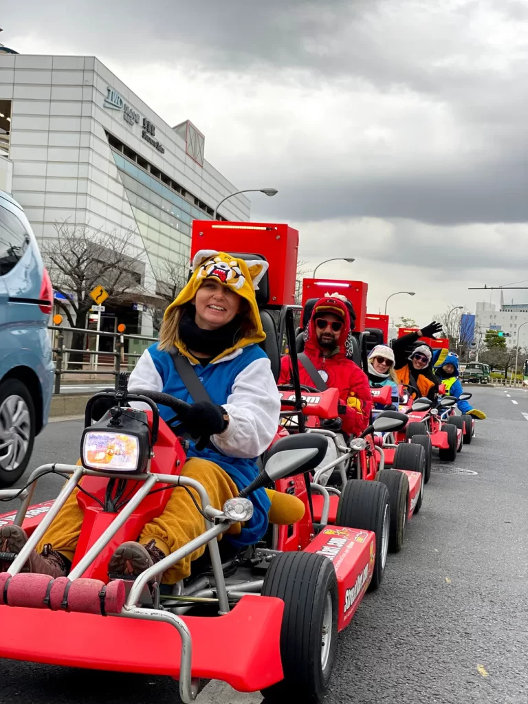 People in a line of red go-kart style parade carts driving down a city street; foreground rider wears a tiger hoodie and smiles at the camera.