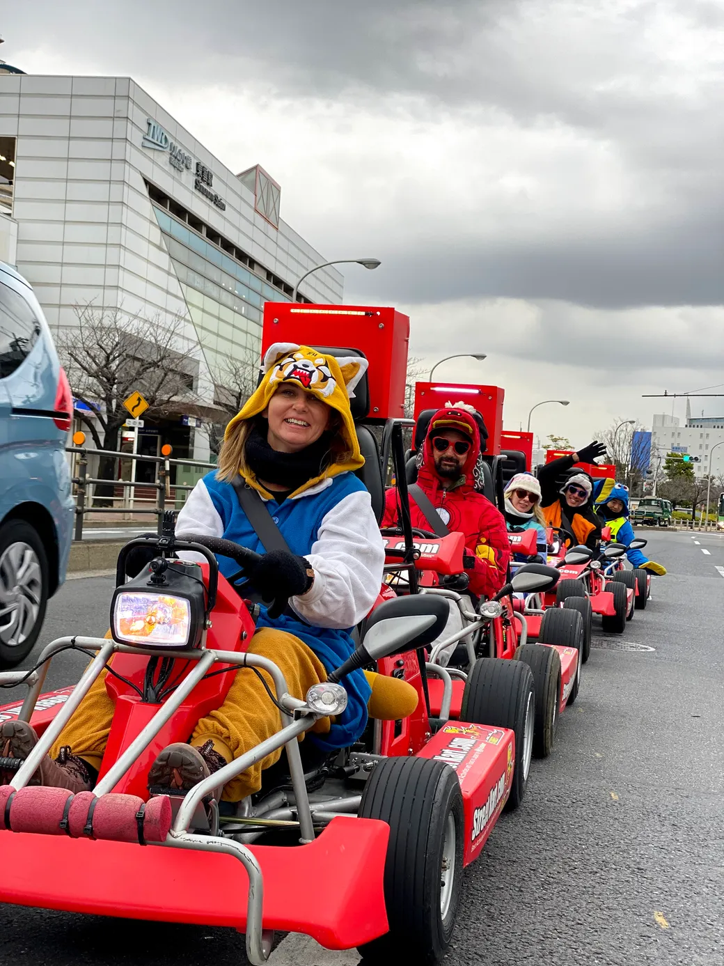 People in a line of red go-kart style parade carts driving down a city street; foreground rider wears a tiger hoodie and smiles at the camera.