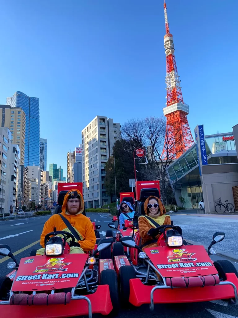 IMG_3639 - StreetKart Three people in orange outfits ride red Street Kart go-karts down a city street, with Tokyo Tower and tall buildings in the background.