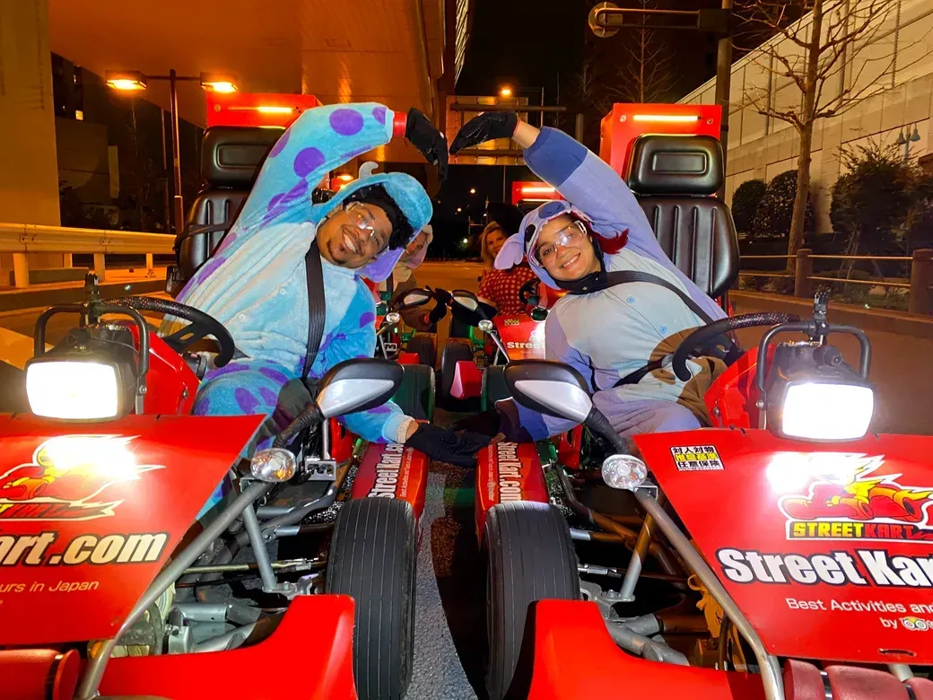 Two people in red Go-Karts at night form a heart shape with their joined arms, wearing colorful cosplay outfits and safety goggles.